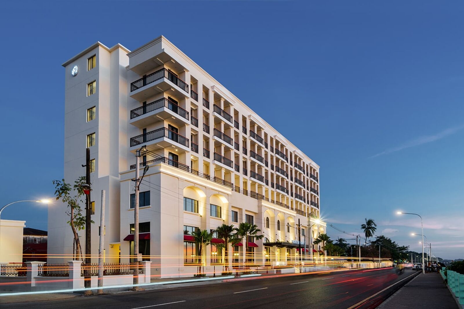 Hotel Suggati seen from the street at dusk, with lit balconies and palm trees